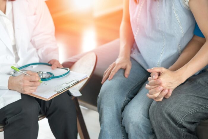 Un couple se tenant la main lors d’une consultation en PMA avec un médecin, qui prend des notes sur un dossier médical avec un stéthoscope à proximité.